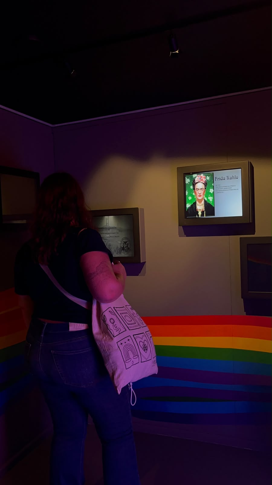 Visitor with a tote bag looking at a Frida Kahlo screen in a museum room with a rainbow on the wall